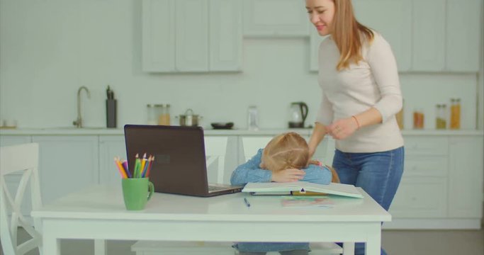 Affectionate caring mother caressing and kissing her cute sleepy tired schoolgirl while daughter sleeping on textbooks at table during doing homework for elementary school in domestic kitchen.