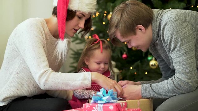 Happy family, parents showing Ney Year decorations to their cute child in front of Christmas tree. Mother in Santa hat, father, and baby girl with Christmas toys and presents near beautiful spruce.