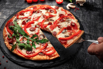 A slice of pizza on a spatula in hand with smoked sausages, cheese, mushrooms, cherry tomatoes,...
