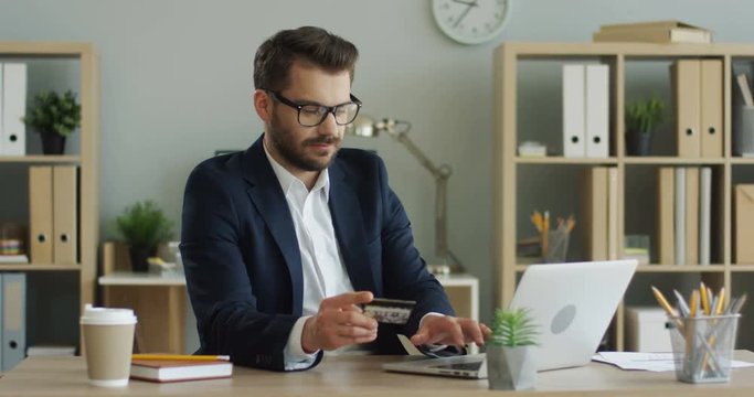 Young attractive man in business style sitting in his cabinet at the desk and buying online on the laptop computer with a credit card and doing yes gesture.