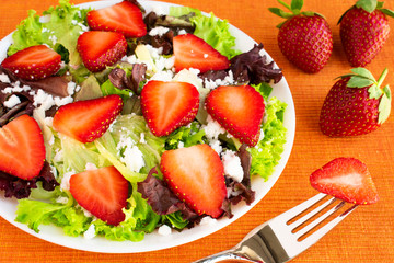 Vegetarian delicious fresh salad with purple and green lettuce leaves, strawberries and goat cheese on orange tablecloth.