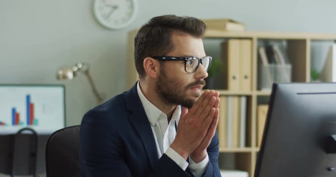 Portrait of the handsome young man in glasses and suit working on the computer in the office room and putting his hands together like praying his work being successful. Close up.