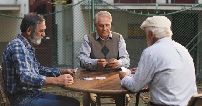 Senior Caucasian man on retirement playing cards with his two best old friends neighbors in the yard on the fresh air. Outdoors.