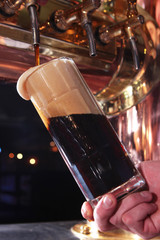 The bartender pours beer in a beer glass. Dark beer, copper equipment for pouring beer. Hand bartender with a mug of beer, foam flows from the mug.