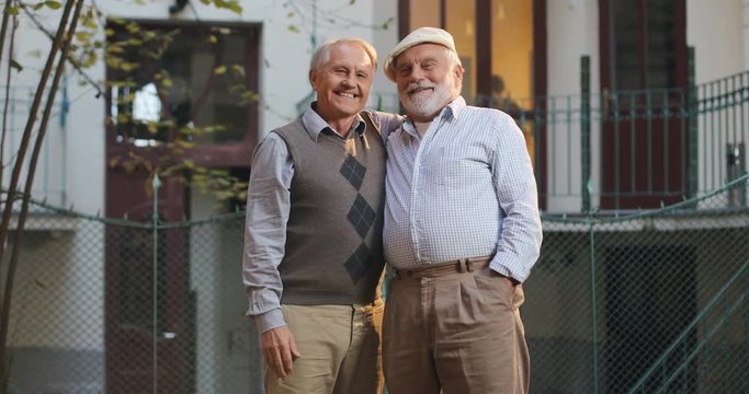 Two Caucasian senior retired men neighbors meeting at the street, greeting, hugging and then posing with smiles to the camera. Outdoor. Portrait.