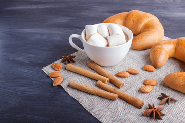 a cup of hot chocolate with marshmallow, croissants, almonds and spices on black wooden background.