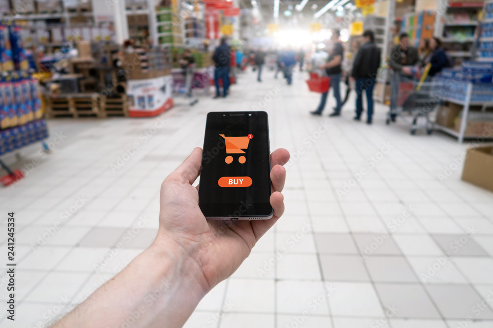 Wall mural hand holding mobile phone on supermarket blur background. shopping basket on a mobile phone screen.