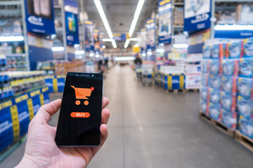 Hand holding mobile phone on Supermarket blur background. Shopping basket on a mobile phone screen. 