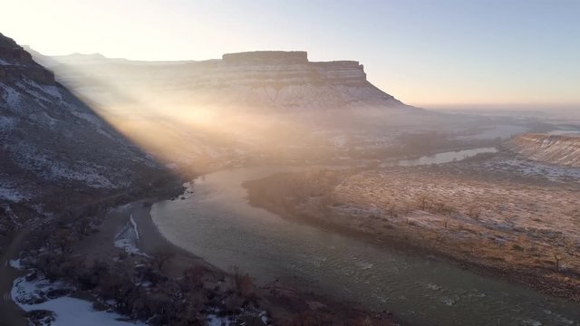 Flying over river as the sun rays shine over the desert landscape in winter as the sun peaks out from behind a cliff in Utah.