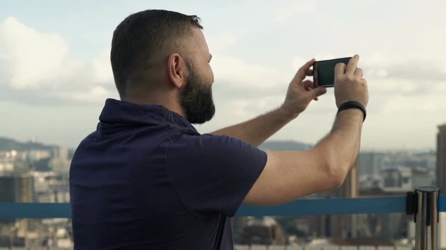 Young man taking photos of cityscape view with cellphone in rooftop bar