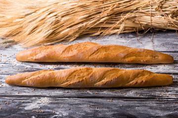 Two bread baguette lying on a wooden table with flour scattered on the background of wheat ears