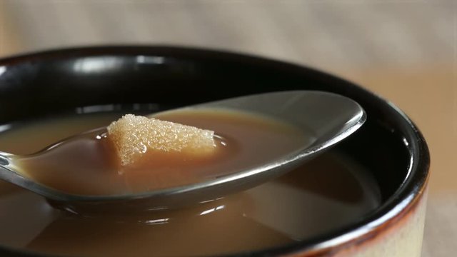 Sugar cube dissolves at rapid speed in tea spoon of coffee with cream. 4K UHD macro, cup edge visible