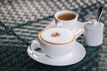 Hot cappuccino in a cup with hot tea and sugar powder in a small jar. Drinks that help refresh the body in the morning. It has a mixture of milk and creamer. Soft focus and blur.