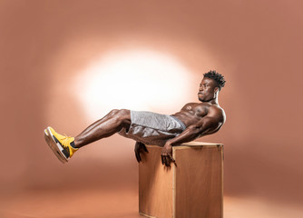 Muscular African American Black athletic man does an ab crunch exercise on the edge of a box in studio with dramatic lighting with a brown background  