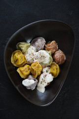 Black bowl with boiled multicolored dumplings or pelmeni, flatlay on a black stone surface, vertical shot