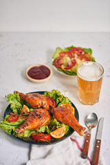 Grilled chicken legs roasted on the grill on dark plate with tomato sauce in a bowl and lettuce leaves, glass mug of beer