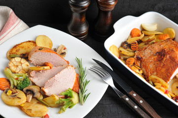 Pork ham in marinade. Around the ham lined vegetables cooked on the grill. Next to it is a plate with slices of pork ham and vegetables. Dark background. Close-up.