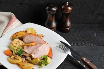 Slices of pork ham with stewed vegetables. On the plate, cutlery. Free space under the text. Dark background. Close-up. 