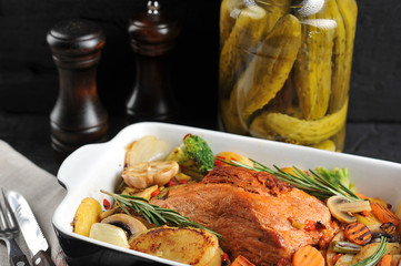 Pork ham in marinade. Around the ham lined vegetables cooked on the grill. In the frame of cutlery and a jar of pickles. Dark background. Close-up.