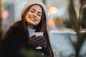 Pretty woman stand on the street in cloudy weather with cup of hot coffee, drink it and smile to camera while talk at phone with copy space background. - Image