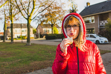 Fototapeta premium Young attractive girl in red winter clothes standing on a street and eating with pleasure her sandwich.