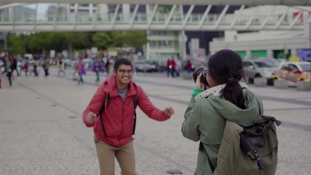 Joyful young man in eyeglasses jumping during photo session outdoor. Young female tourist taking pictures of her boyfriend with professional photo camera. Travel concept
