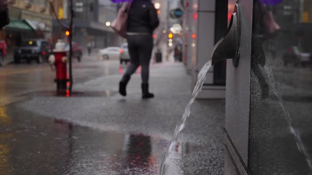 A slow motion view of water flowing from a city building's industrial downspout as pedestrians pass by on the sidewalk nearby. Shot at 60fps.  	