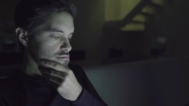 Sleepy man rubbing face and working at night. Tired young man sitting on couch and using digital device in dark room. Networking concept