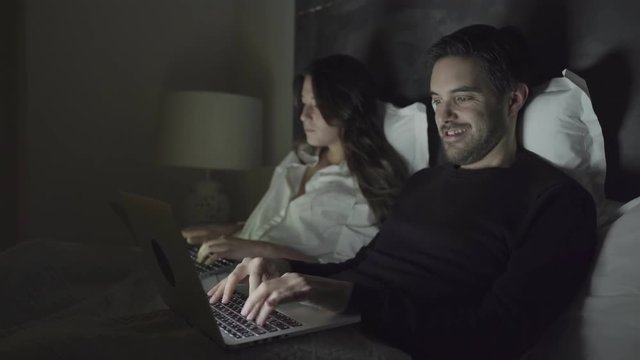 Couple using laptops in bedroom at night. Beautiful focused girl and smiling man typing on laptops together in bed. Technology concept
