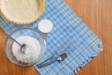 Top view of the ingredients of a coconut cream pie on a blue plaid placemat on wooden table including flaked coconut, sugar, eggs and the pie shell. Copy space