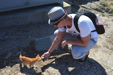 Caucasian man with a backpack crouched down and holding an action camera in his outstretched hand. Red kitten sniffs the camera