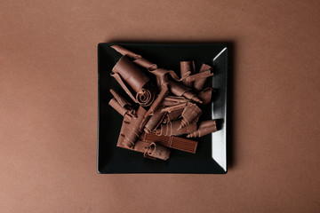 Plate with chocolate curls and pieces on color background, top view