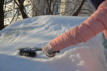 Winter portrait of a young woman cleaning snow from a car. Beauty blonde Model Girl laughs and...