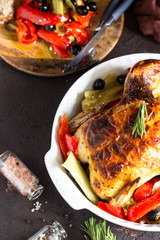 Tasty food: whole roasted chicken with vegetables (red bell pepper, leek  and olives) in a baking dish on a old brown background. 