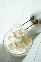 Whipped cream in a bowl and electric hand mixer on bright table.