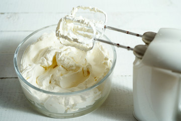 Whipped cream in a bowl and electric hand mixer on bright table.
