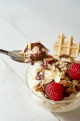 Bowl with dessert with whipped cream, raspberries and nuts with spoon on white wooden table.