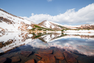 Spring mountain lake with clear water and red stones. Picturesque winter landscape with snowy hills under a blue sky