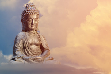 Buddha statue sitting in meditation pose against sunset sky with golden tones clouds.