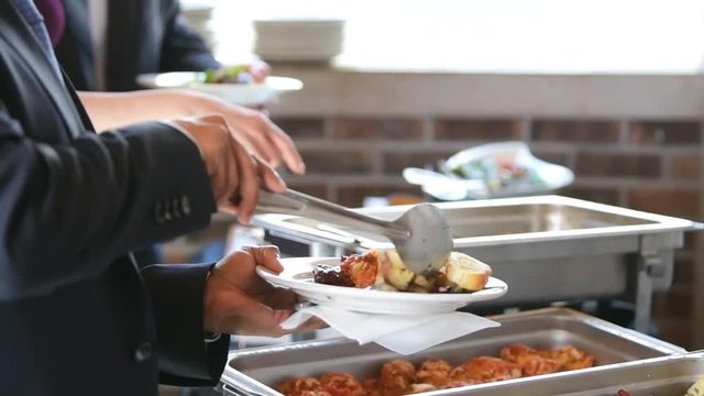 Slow motion, closeup of side profile of people taking, serving food on plate using spoon from hot buffet tray with fresh roasted potatoes, chicken in banquet, wedding, or restaurant