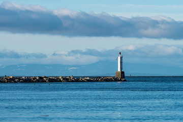 small light house at the end of rocky shore line with drift woods on the river with snow covered mountains in the background over the horizon under cloudy blue sky.
