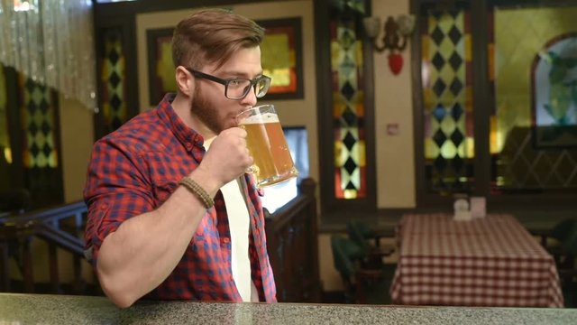 Portrait of a bearded handsome man in a red checkered shirt with glasses and a glass of beer in his hands against the background of a pub.