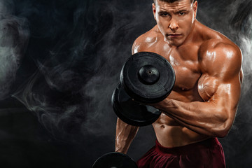 Brutal strong muscular bodybuilder athletic man pumping up muscles with dumbbell on black...