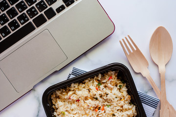 Delicious fried rice in lunch box on napery with wooden spoon and fork and computer laptop at...