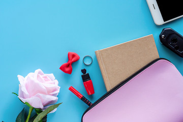 Top view of pink lady bag with diary book, pen, lipstick, bow, ring, car key, smartphone and sweet rose on blue background for women accessories and lady lifestyle concept