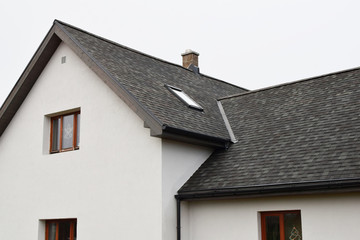 Obraz premium Part of white house with new wooden eaves, plastic rain gutter, window and chimney in grey shingles roof and brown window in facade with copy space on sky.