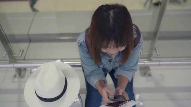 Happy Asian woman using and checking her smartphone while sitting on chair in terminal hall while waiting her flight at the departure gate in international airport. Women happy in airport concept.