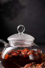 A glass teapot with a freshly brewed vitamin rosehip drink and dry fruits on a decorative board. Rosehip tea. Vitamin drink. Dark background. Copy space.