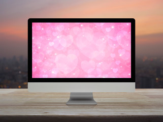 Fototapeta premium Desktop modern computer monitor with pink love heart screen on wooden table over blur of cityscape on warm light sundown, Business internet dating online, Valentines day concept