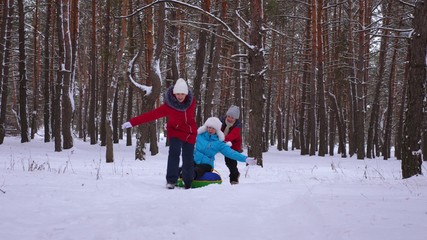 Fototapeta na wymiar funny children ride their mom on sled and an inflatable snow tube in pine forest. happy family mom and kids playing in winter park and forest for christmas holidays.